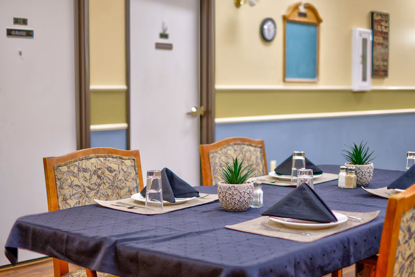 A dining table set with a dark tablecloth, beige placemats, black folded napkins on white plates, clear drinking glasses, salt and pepper shakers, and small potted plants. The background shows a hallway with closed doors and a wall clock.