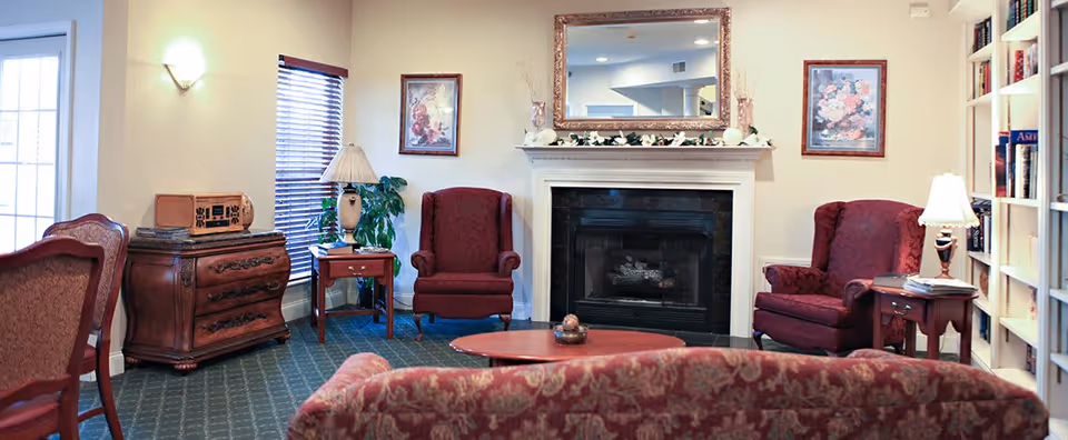 A cozy living room with patterned red upholstered chairs and sofa arranged around a wooden coffee table. There is a fireplace with a white mantel decorated with flowers and a large mirror above it. The room also features a wooden side table with a lamp, a chest of drawers with a vintage radio, framed floral artwork on the walls, a window with blinds, and a bookshelf filled with books.
