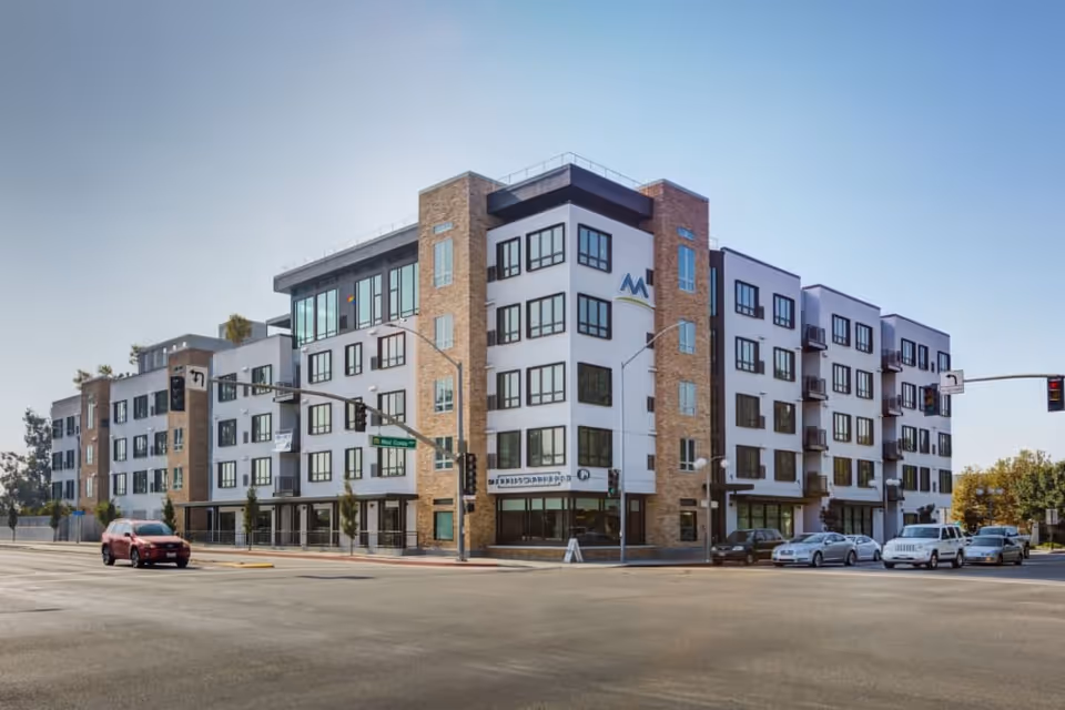 Modern multi-story senior living building with brick and white facade on a street corner with parked cars.