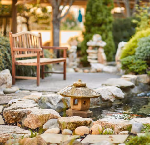 A peaceful outdoor garden area featuring a wooden bench, a small stone lantern, a pond surrounded by rocks, and various green shrubs and trees in the background.
