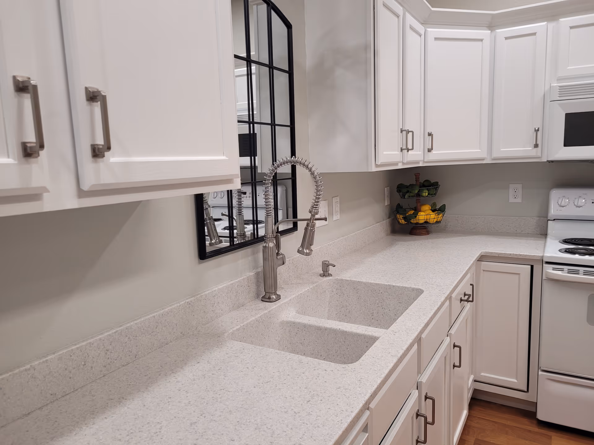A modern kitchen with white cabinets and drawers, a double sink with a flexible spring faucet, a white electric stove, a microwave, and a two-tier fruit basket filled with lemons and limes on the countertop.