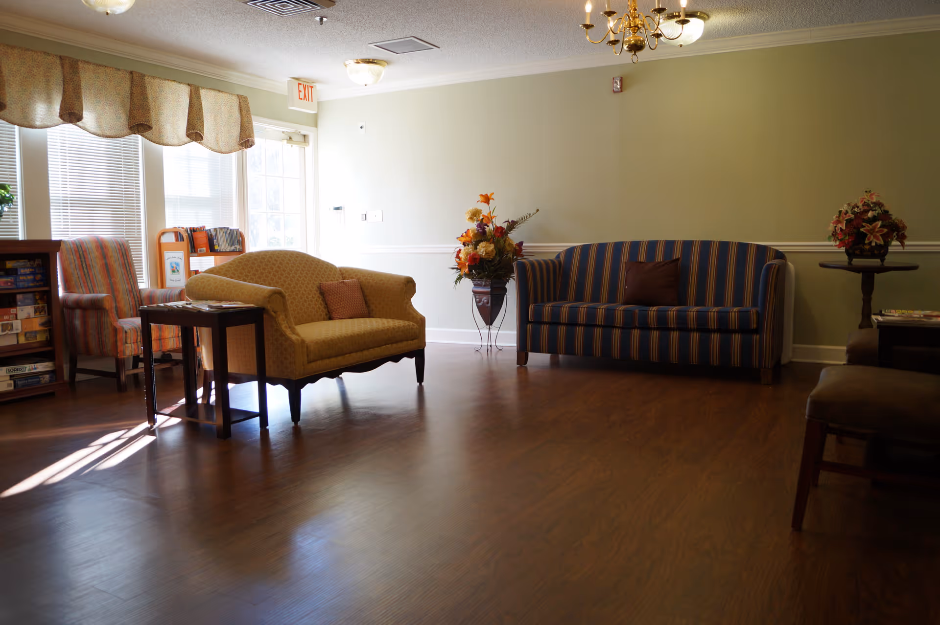 A bright senior living common room with upholstered sofas and chairs, floral arrangements, and large windows letting in sunlight.