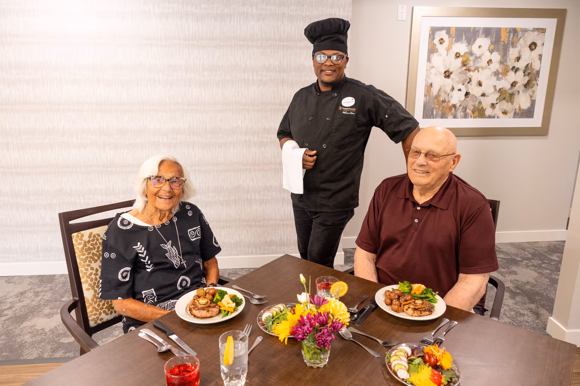 Two elderly residents sit at a dining table with plates of food while a chef stands behind them smiling.