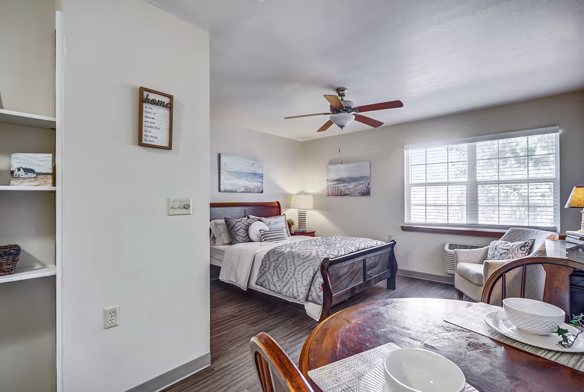 Bright studio-style bedroom with a bed, ceiling fan, window, armchair, and a small dining table in the foreground.