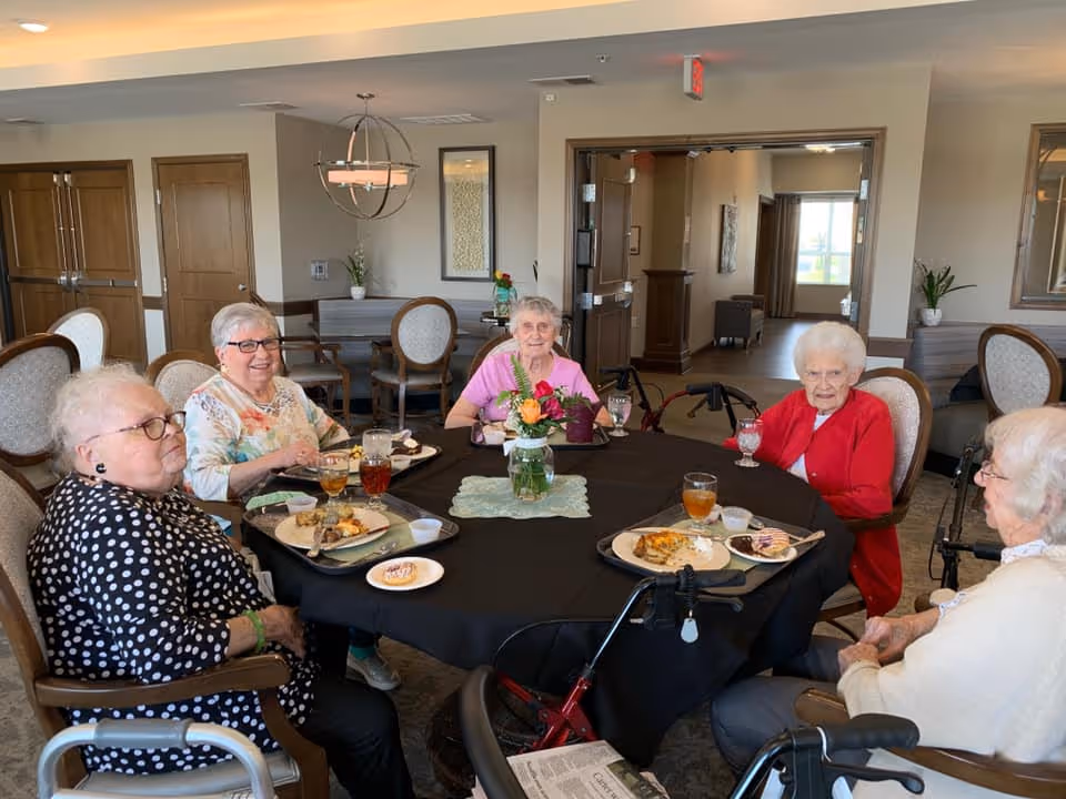 Five elderly women sitting around a round dining table with black tablecloth, enjoying a meal together in a well-lit dining room. The table has plates with food, glasses of iced tea, and a floral centerpiece. The room has wooden doors, chairs with cushioned seats, and a chandelier hanging from the ceiling.