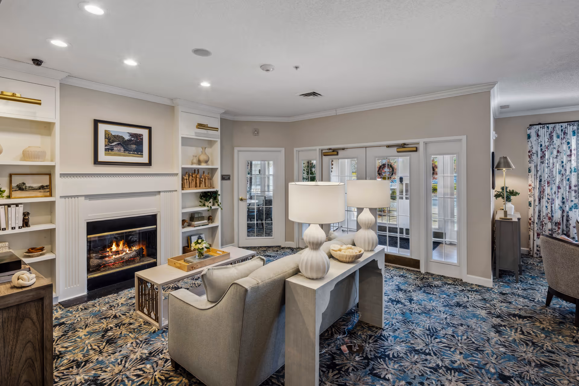 A cozy living room area with a lit fireplace surrounded by built-in shelves holding decorative items and framed pictures. In front of the fireplace is a light gray sofa with a console table behind it, topped with two white lamps and a bowl. The room has patterned carpet, a glass door leading outside, and floral curtains on the right side.