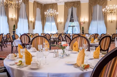 Elegant dining room with round tables set with white tablecloths, yellow folded napkins, glassware, and floral centerpieces. The room features large windows with draped curtains, chandeliers, and striped upholstered chairs.
