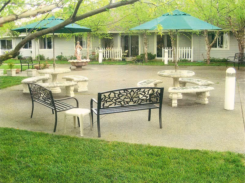 Outdoor patio area with two black metal benches featuring decorative backs, two round stone tables with curved stone benches, green umbrellas providing shade, a small white stool, and a fountain statue in the background near a white picket fence and residential building.