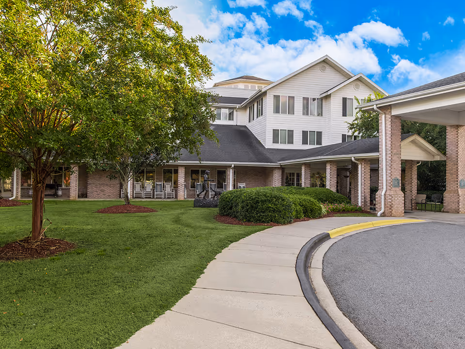 Exterior view of Solista Hickory senior living facility showing a multi-story building with white siding and brick accents. There is a covered entrance with brick columns, a curved driveway, green lawn, trees, and bushes under a partly cloudy blue sky.