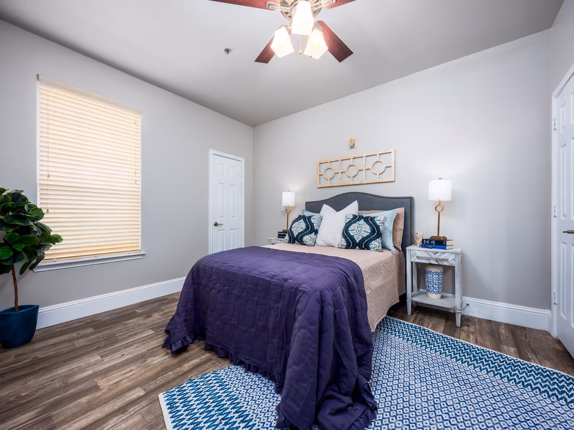 A neatly arranged bedroom with a bed covered in a purple blanket and multiple decorative pillows. The room features wooden flooring, a blue and white patterned rug, two bedside tables with lamps, a potted plant near a window with closed blinds, and a ceiling fan with lights.