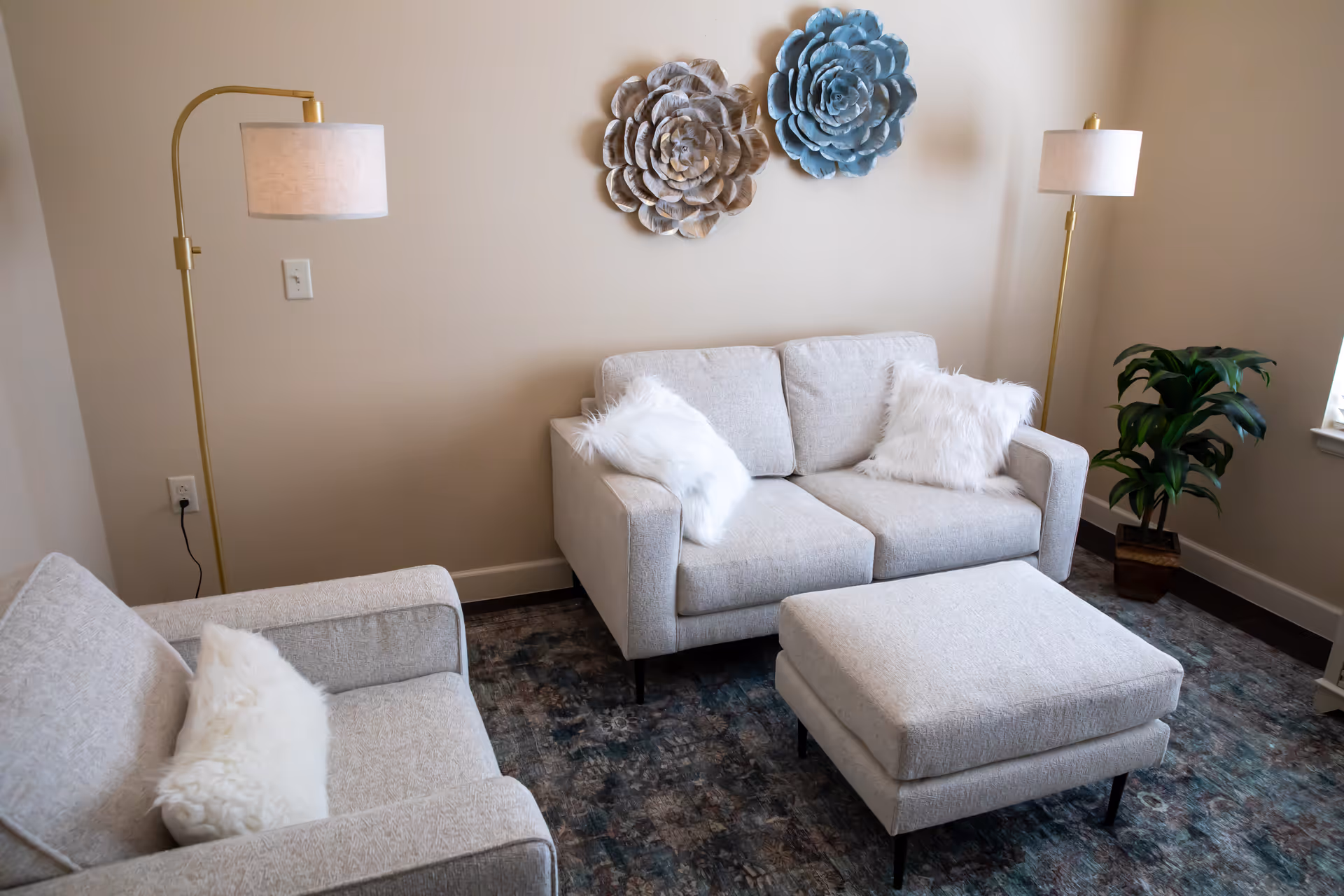 A cozy living room with a beige loveseat and armchair, both adorned with white fluffy pillows. A matching ottoman sits in front of the loveseat on a patterned rug. Two decorative flower wall hangings are mounted on the beige wall behind the loveseat. Two tall floor lamps with white shades flank the seating area, and a green potted plant is placed near a window on the right.