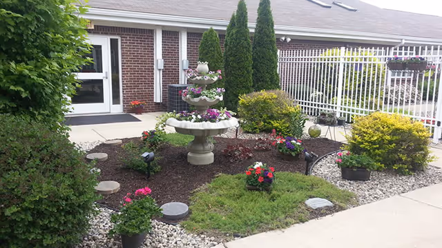 Small landscaped courtyard with a tiered fountain, colorful flower beds, shrubs, and a white fence in front of a brick building entrance.