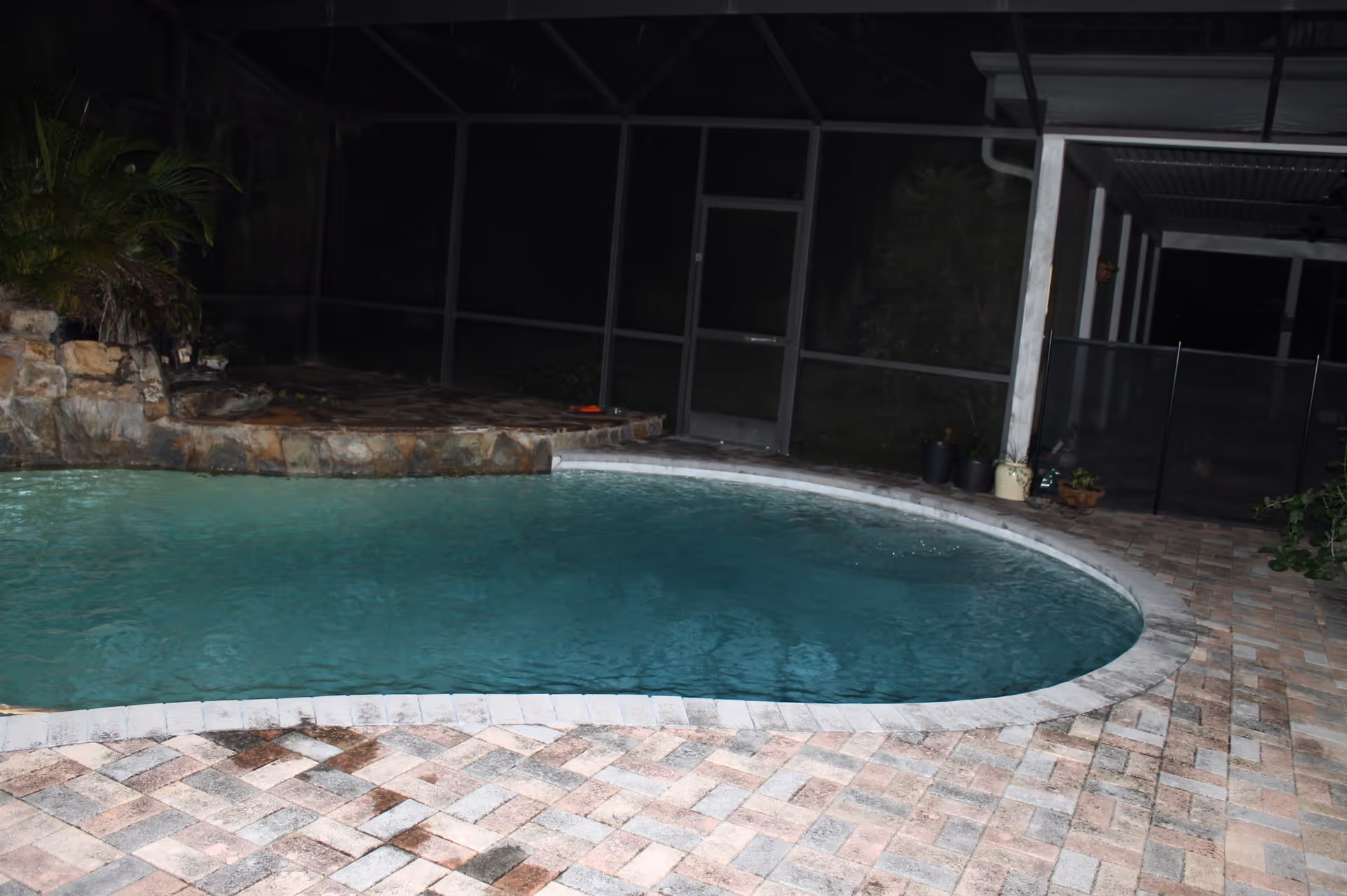 Screened-in backyard swimming pool with a stone waterfall and brick paver deck at night.
