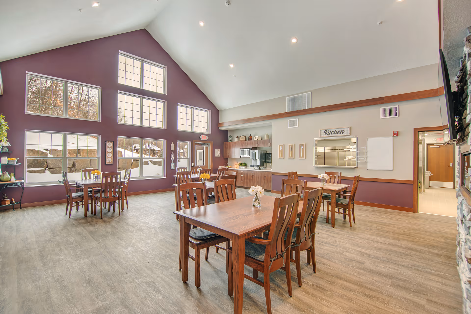 Bright dining room with wooden tables and chairs, a vaulted ceiling, large windows showing snow outside, and a serving window labeled 'Kitchen'.