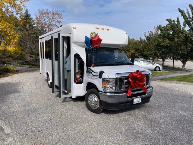 A white shuttle bus decorated with a large red bow on the front grille and three balloons (red, blue, and multicolored) tied near the driver's side mirror. The bus is parked on a paved area with trees and parked cars in the background under a partly cloudy sky.