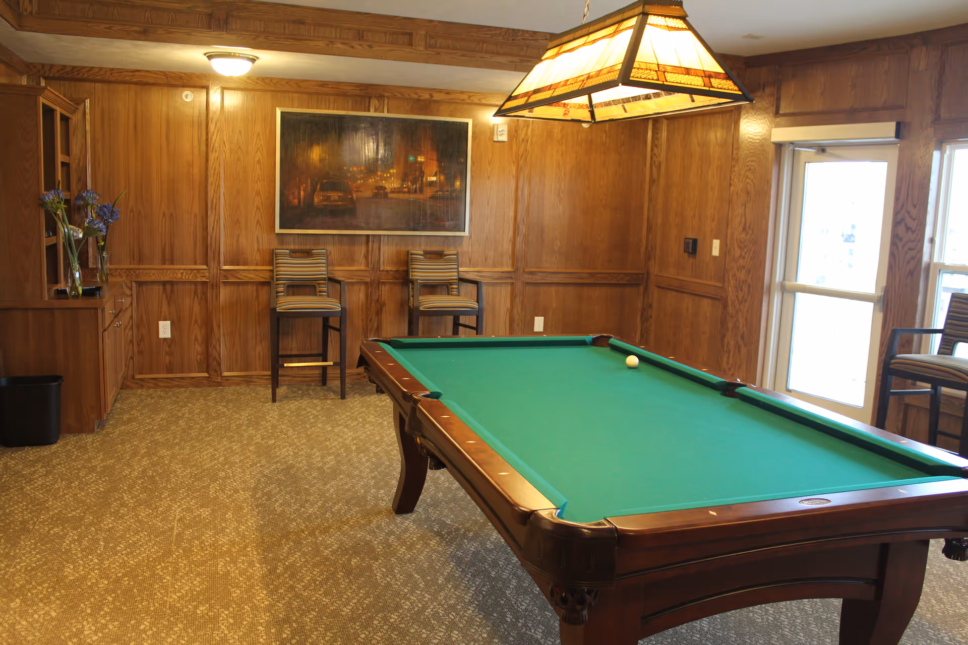 A room with wood-paneled walls featuring a pool table with a green felt surface and a single white ball on it. Two striped chairs are placed against the wall beneath a framed painting. A hanging light fixture with a stained glass design is above the pool table. There is a door and a window on the right side, and a small cabinet with a vase of flowers on the left.