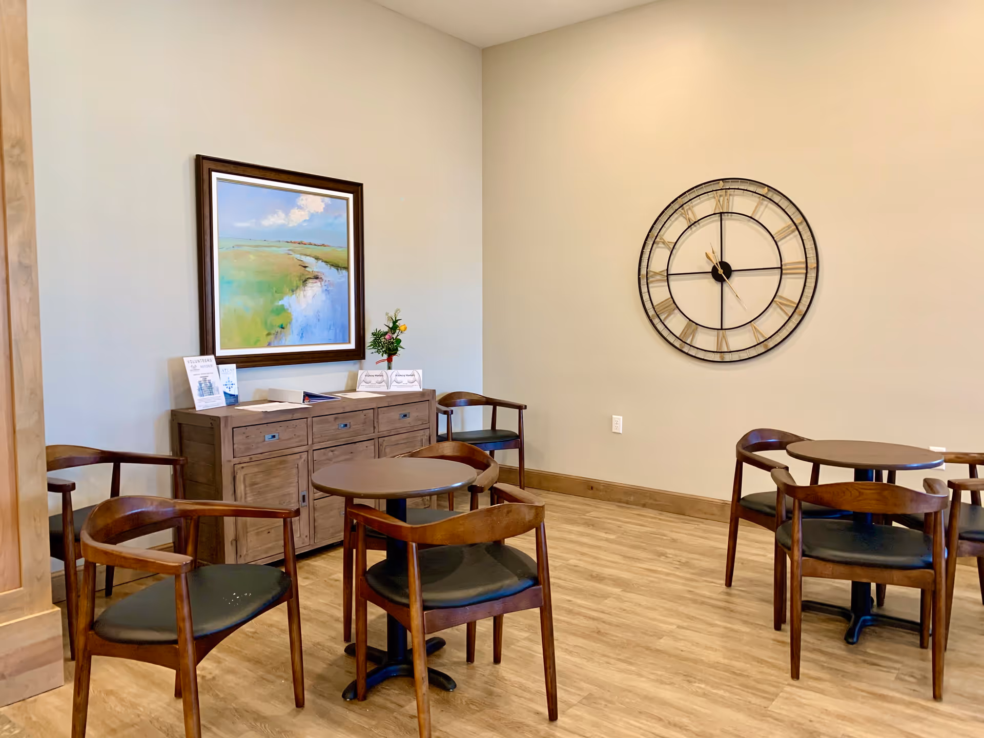 Small communal dining area with round tables and wooden chairs, a sideboard with a painting, and a large wall clock.