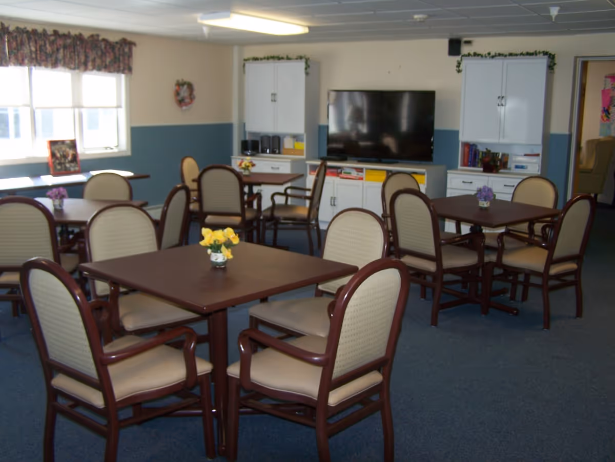 A common area with several square tables surrounded by cushioned chairs. Each table has a small flower arrangement. In the background, there is a large flat-screen TV mounted on a white cabinet with shelves and storage. The walls are painted beige and blue, and there is a window with floral curtains letting in natural light.