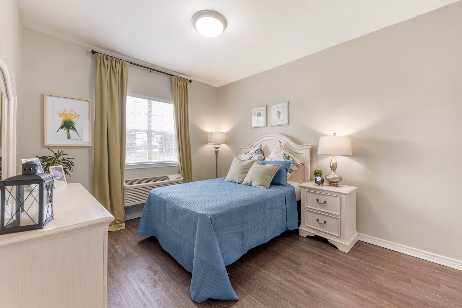 A cozy bedroom with a bed covered in a blue quilt and multiple pillows. There is a white nightstand with a lamp and a small plant on it next to the bed. A window with beige curtains allows natural light into the room. A dresser with decorative items and framed artwork on the walls complete the room.