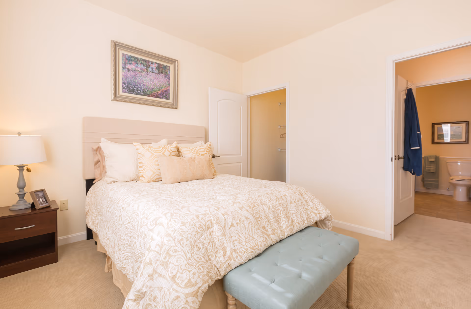 Sunlit bedroom with a patterned bed, bedside table and lamp, and open doors leading to a closet and bathroom.