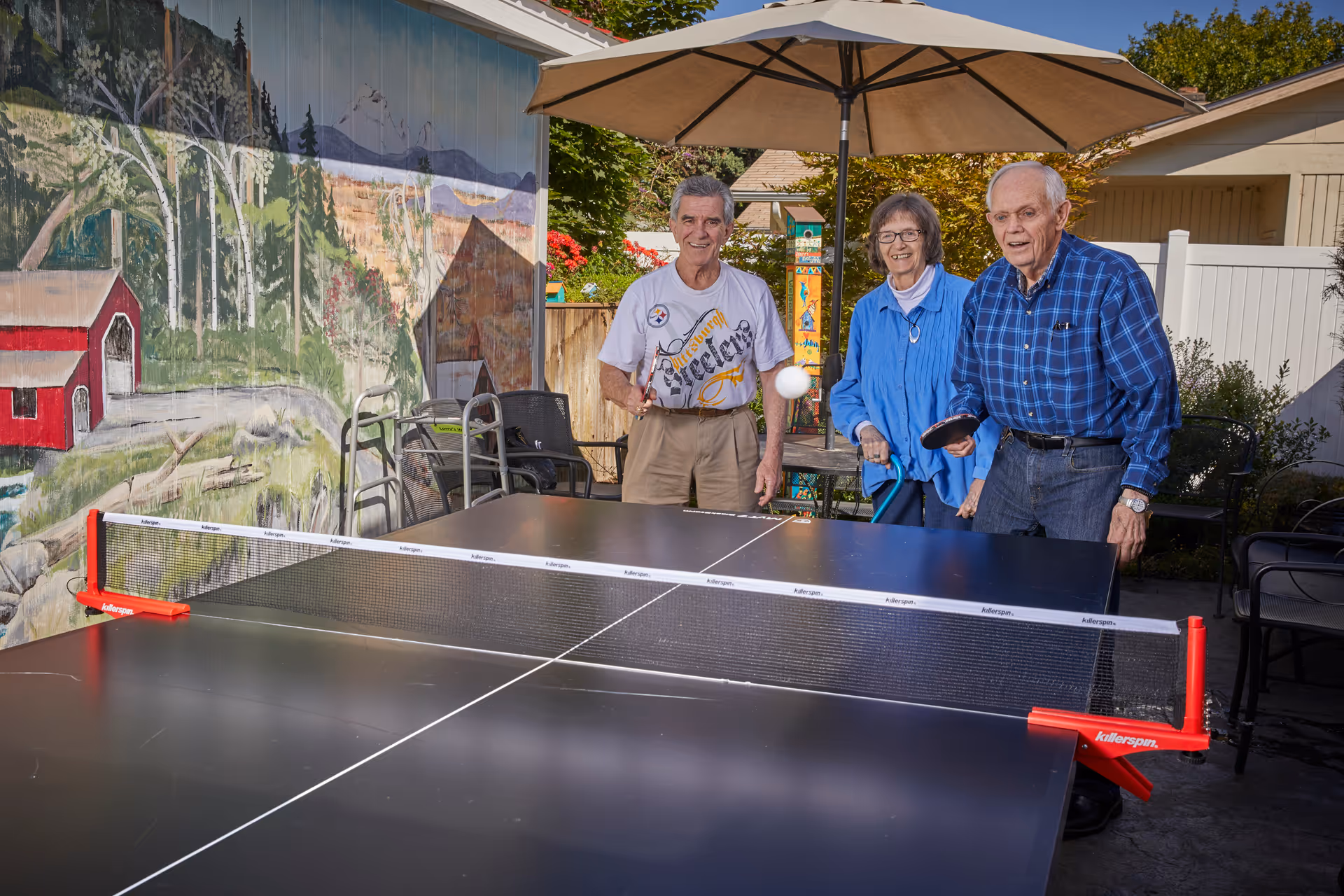 Three elderly people playing table tennis outdoors under a large umbrella. Behind them is a colorful mural depicting a rural scene with trees, a barn, and mountains. The setting appears to be a patio area with chairs and greenery.