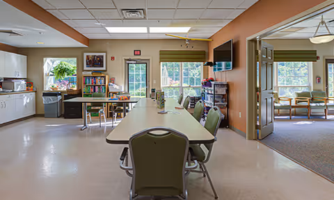 Interior view of a common area in a senior living facility featuring a long table with chairs, a kitchenette with white cabinets and a microwave, a bookshelf with books, a door with a window, and large windows letting in natural light. Adjacent room visible through an open doorway with additional seating and windows.