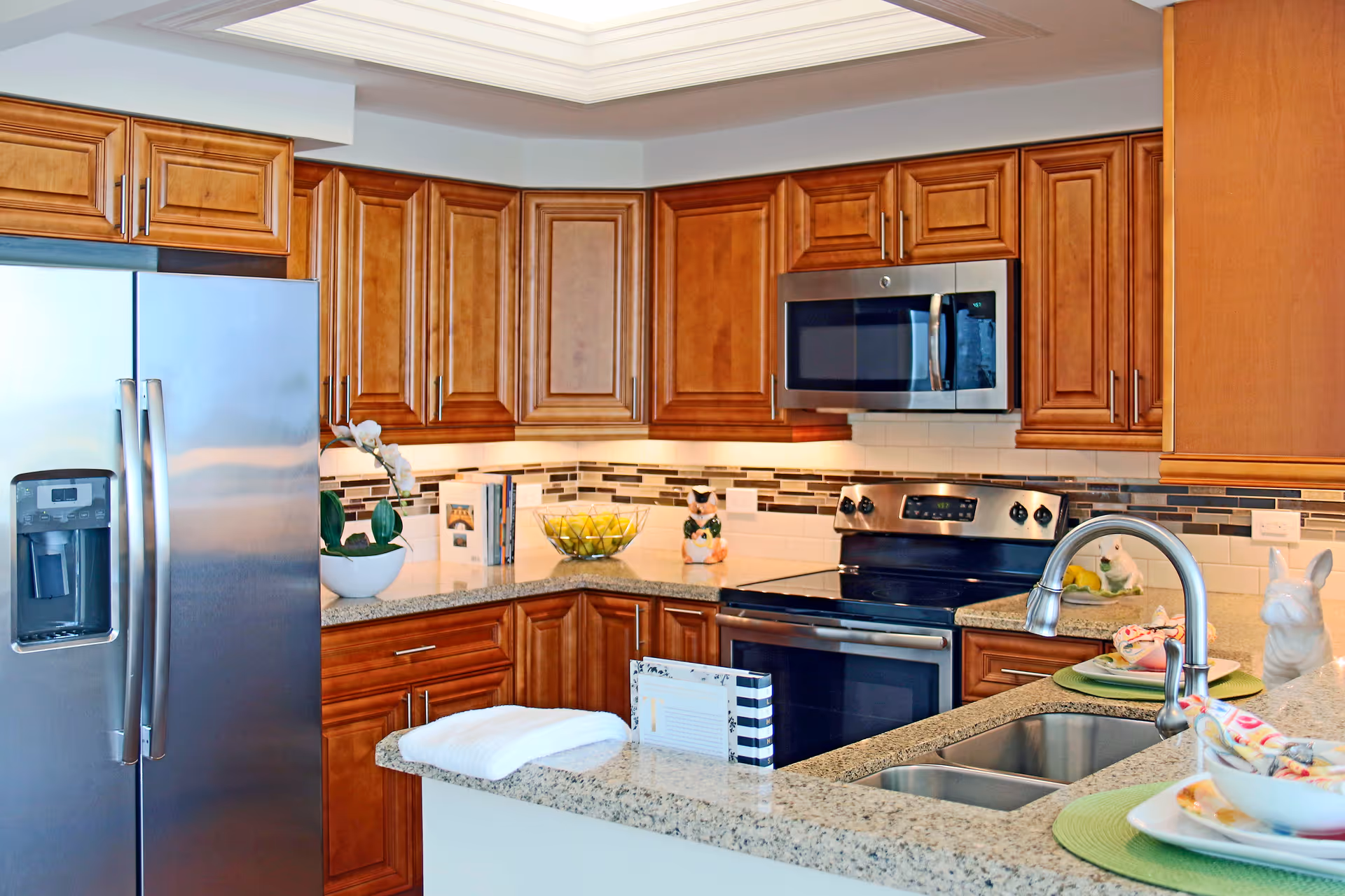 A modern kitchen with wooden cabinets, a stainless steel refrigerator, microwave, and stove. The countertop is granite with a double sink and decorative items including a bowl of lemons, a potted plant, and ceramic figurines.