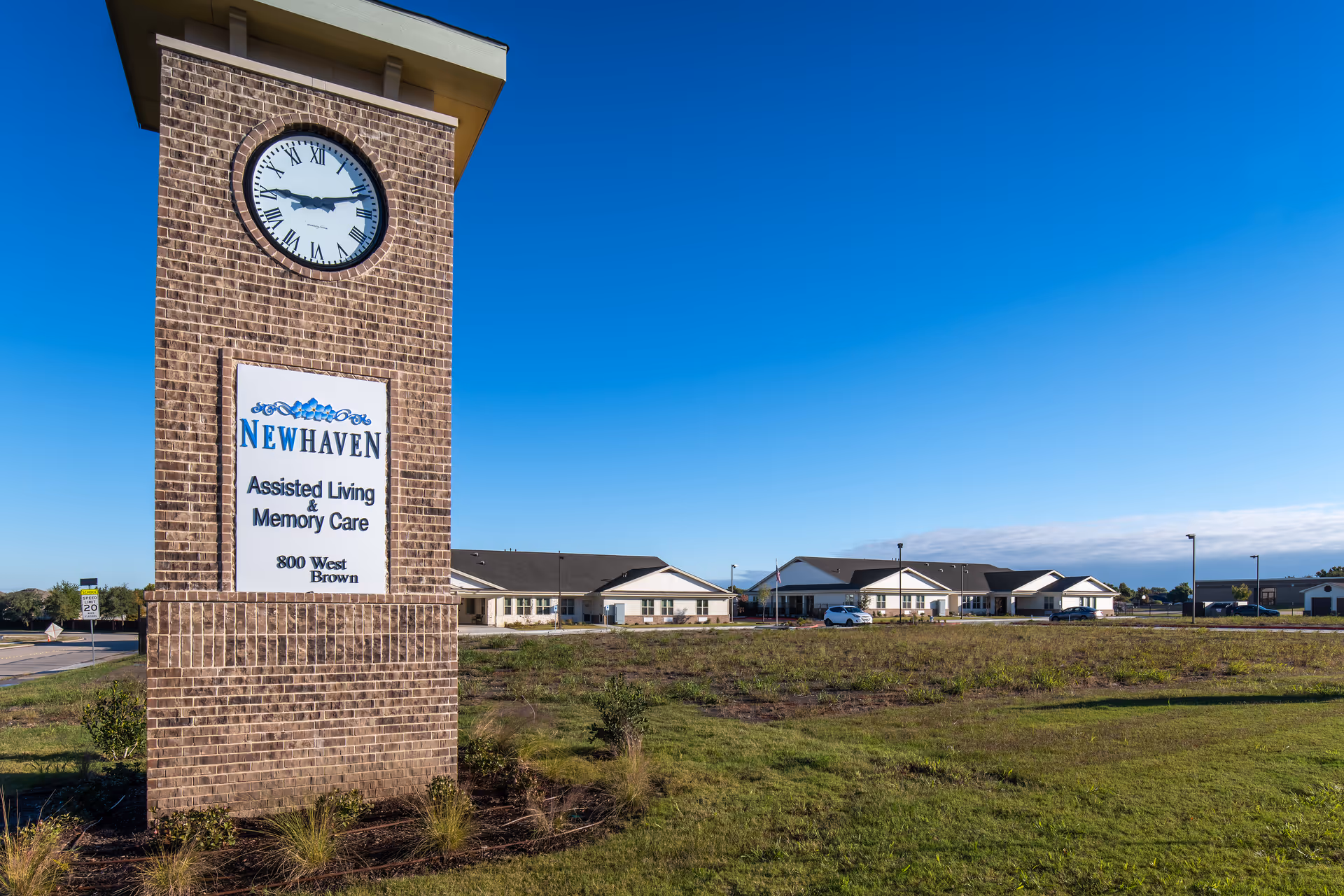 Exterior view of New Haven Assisted Living and Memory Care of Wylie facility with a large brick clock tower sign displaying the facility name and address, surrounded by a grassy area under a clear blue sky.
