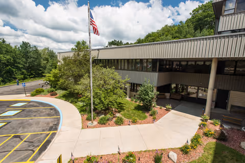 Front entrance of a nursing and rehabilitation center with an American flag, landscaped walkway, and adjacent parking area.