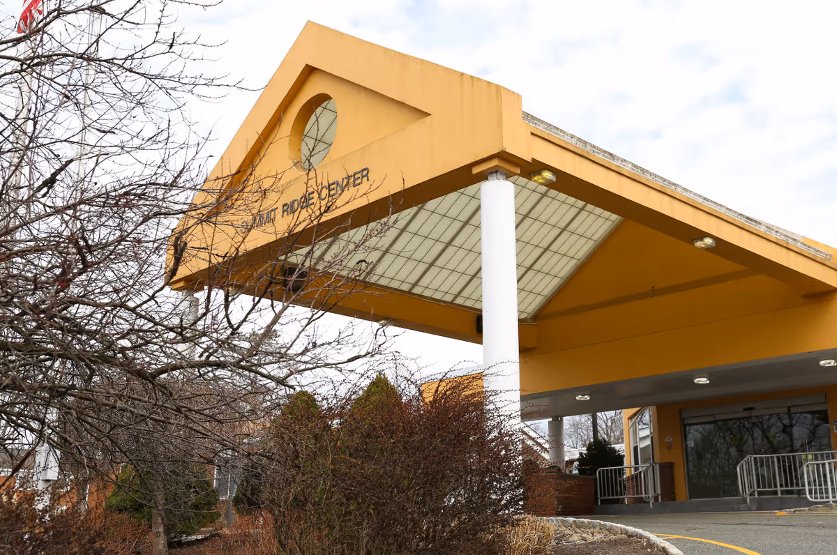 Exterior view of a building entrance with a large yellow canopy supported by white columns. The building has a sign that reads 'Summit Ridge Center'. Leafless trees and bushes are visible in the foreground under a cloudy sky.