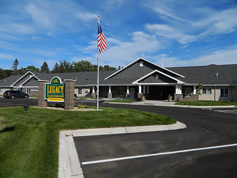 Exterior view of The Legacy Assisted Living building in Morris, MN, showing a single-story structure with a covered entrance, an American flag on a flagpole, a green lawn, and a parking lot in front.
