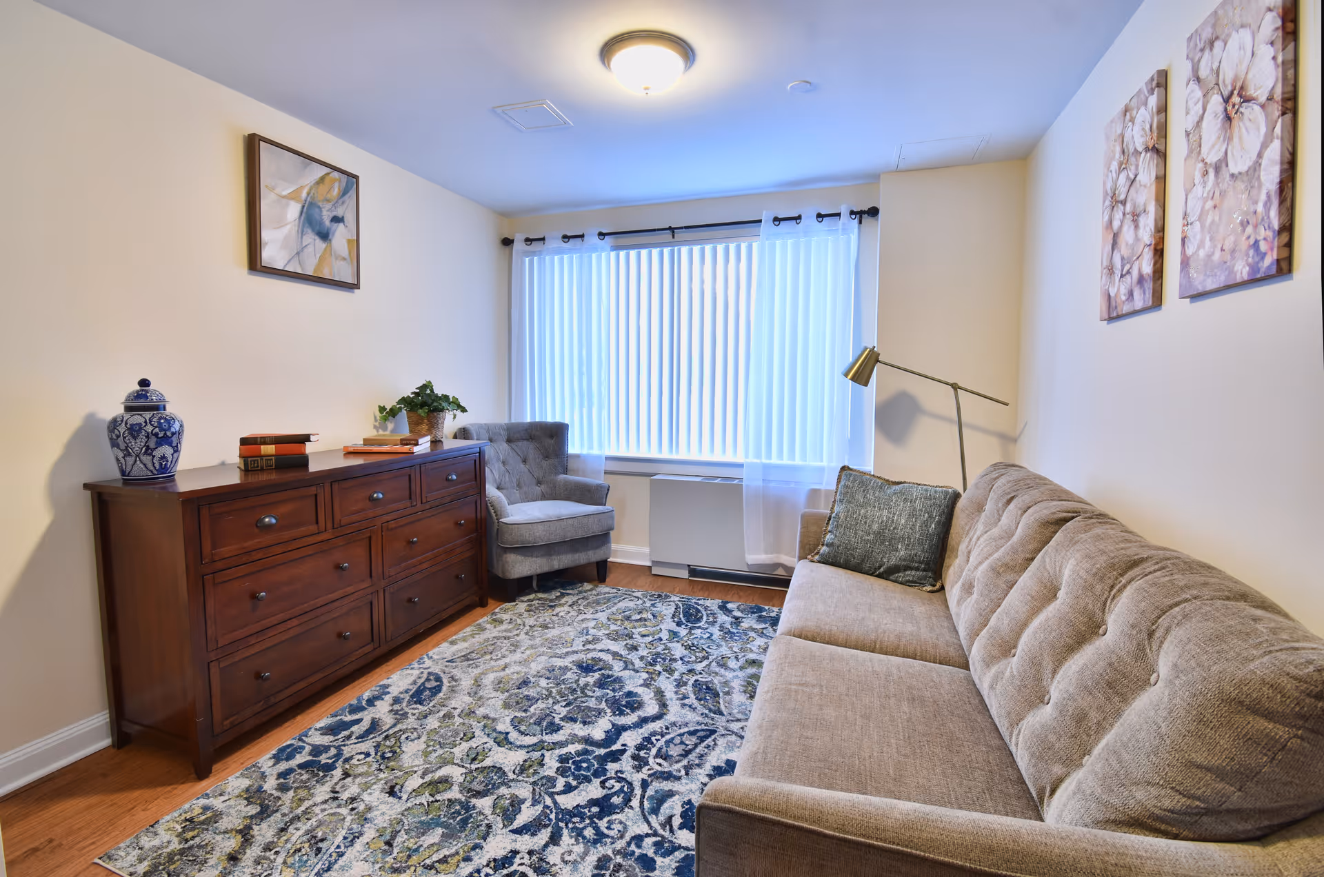A cozy living room with a beige tufted sofa, a gray armchair, a wooden dresser with decorative items including a blue and white vase and books, a patterned area rug, and two floral paintings on the wall. A large window with vertical blinds allows natural light into the room.