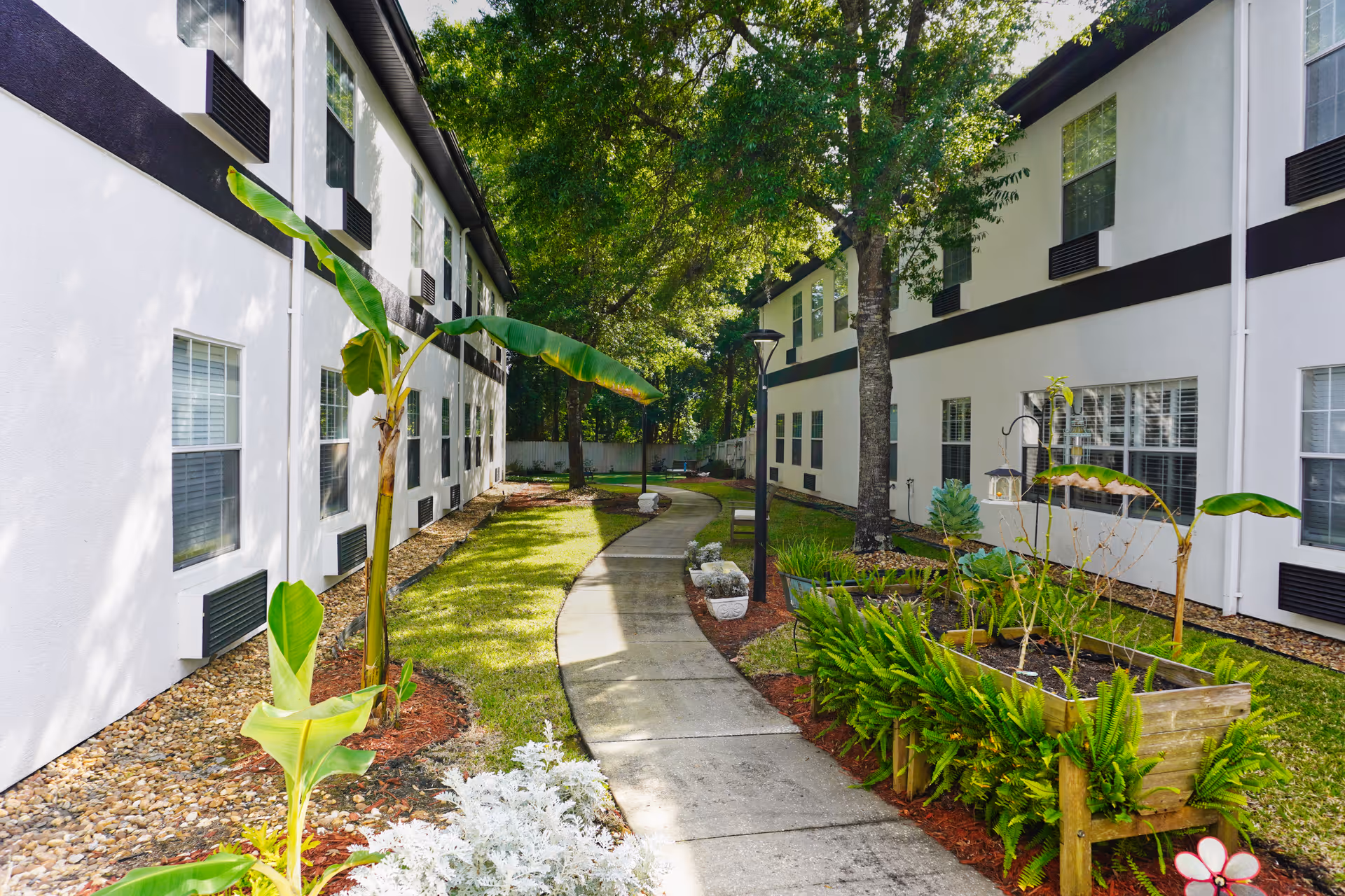 A landscaped courtyard walkway lined with planters and trees between two white residential building wings.