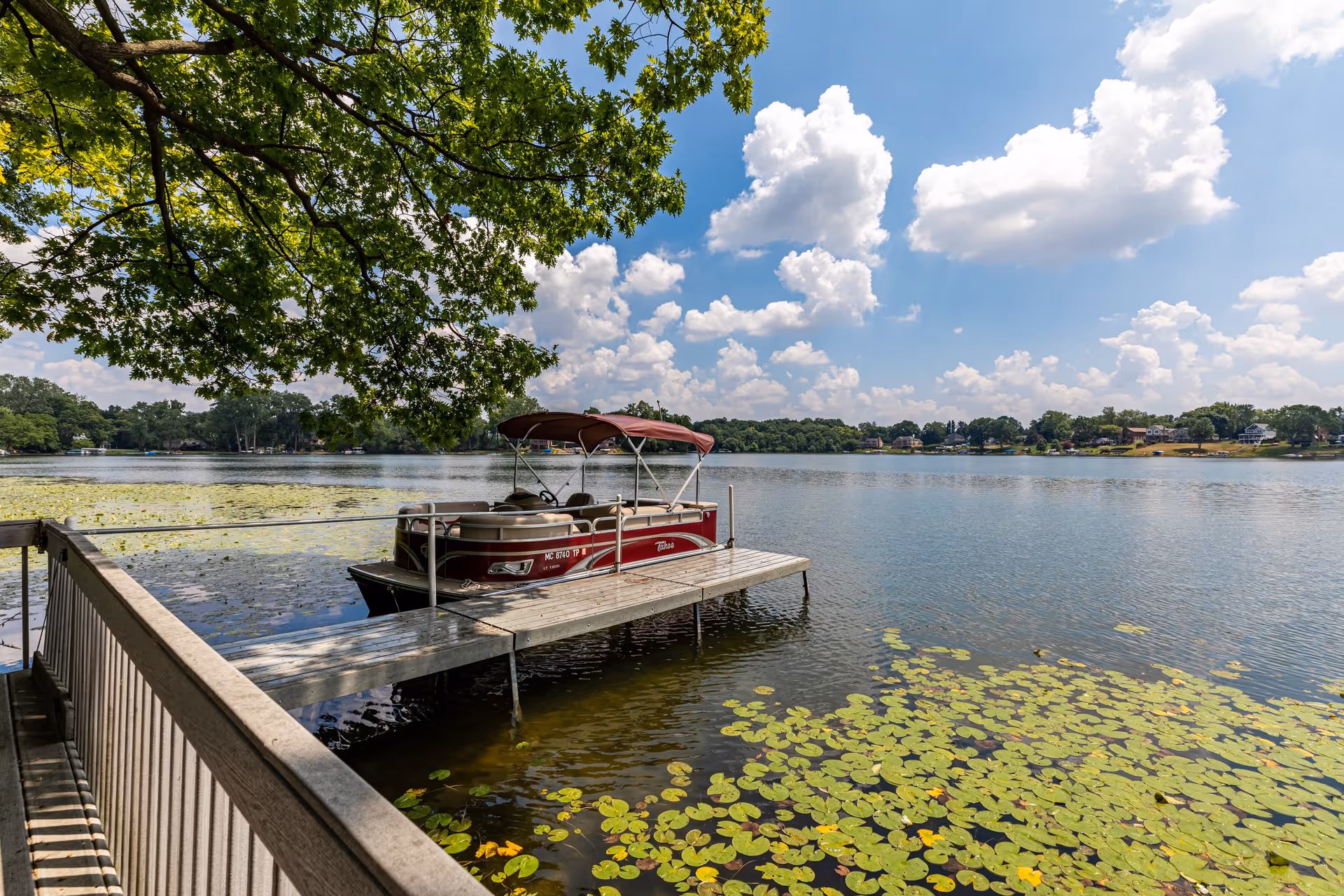 A red pontoon boat docked at a wooden pier on a calm lake surrounded by lily pads. The sky is blue with scattered white clouds, and green tree branches extend over the water from the left side of the image.