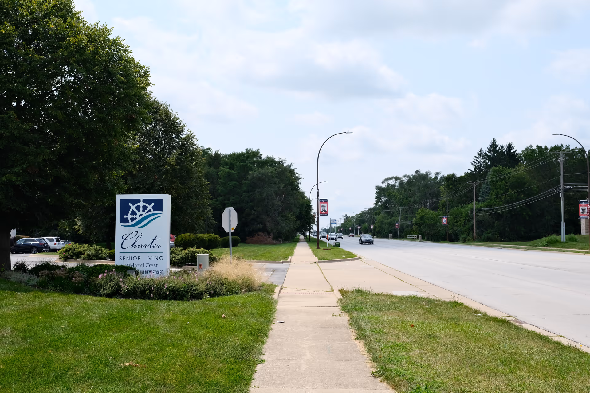 Sidewalk along a street with a sign for Charter Senior Living of Hazel Crest on the left side, surrounded by grass and bushes, with trees and parked cars in the background under a partly cloudy sky.