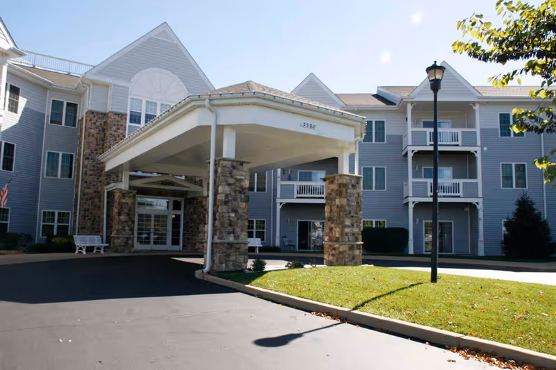 Exterior view of a multi-story senior living facility with gray siding and stone accents. The entrance features a covered drop-off area supported by stone pillars. There are balconies on the upper floors and a well-maintained lawn with a streetlamp in front.
