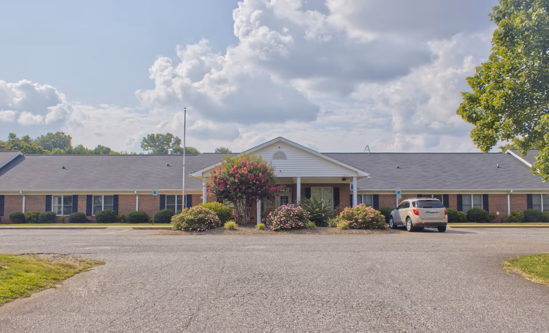 Front exterior view of a single-story brick building with a gray roof, a central entrance with a white portico, and landscaping including bushes and flowering plants. A silver SUV is parked near the entrance, and the sky is partly cloudy.