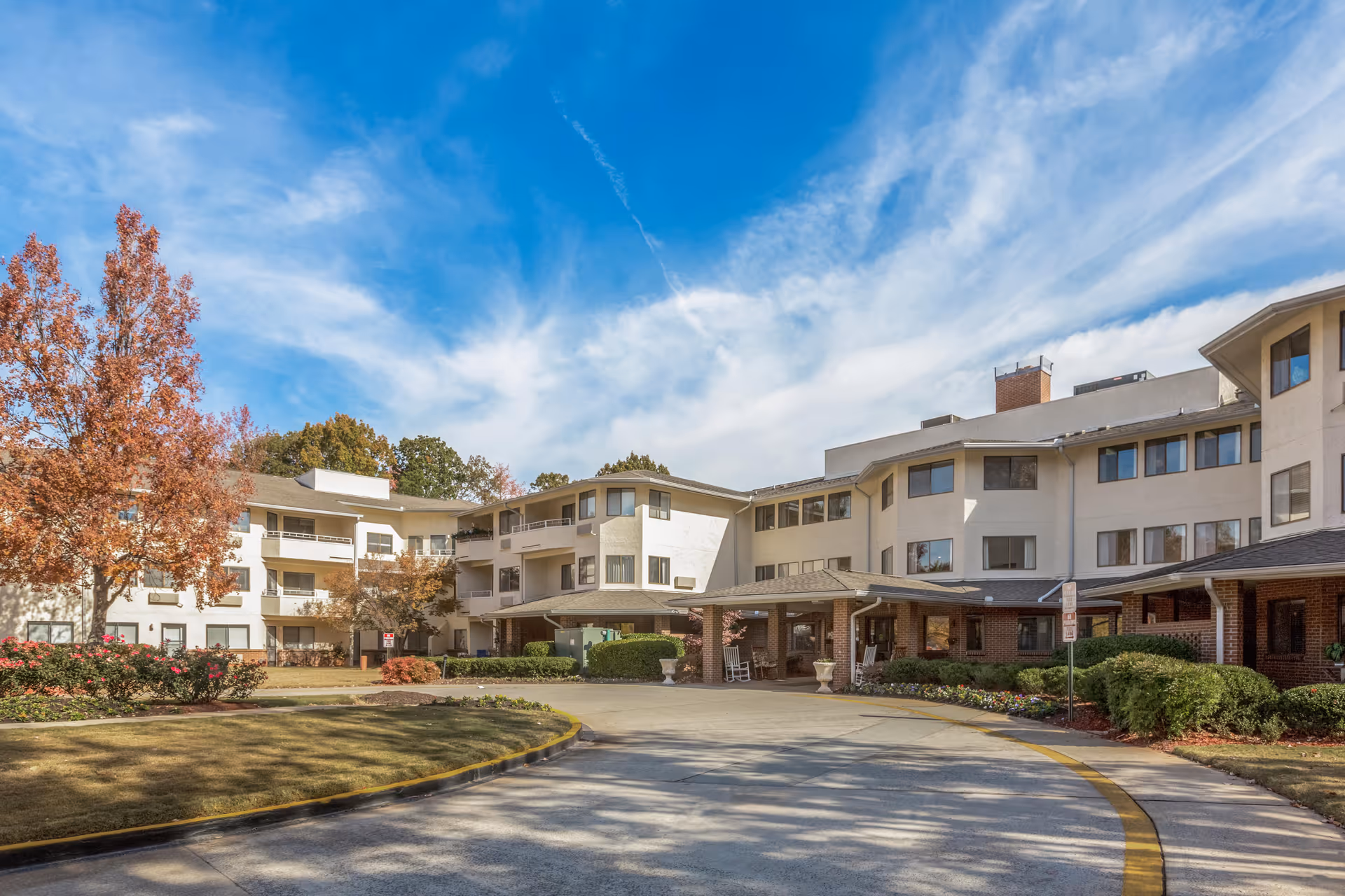 Exterior view of a multi-story senior living facility with beige walls and numerous windows. The building has a covered entrance with rocking chairs and is surrounded by landscaped bushes, flowers, and trees with autumn foliage under a partly cloudy blue sky.