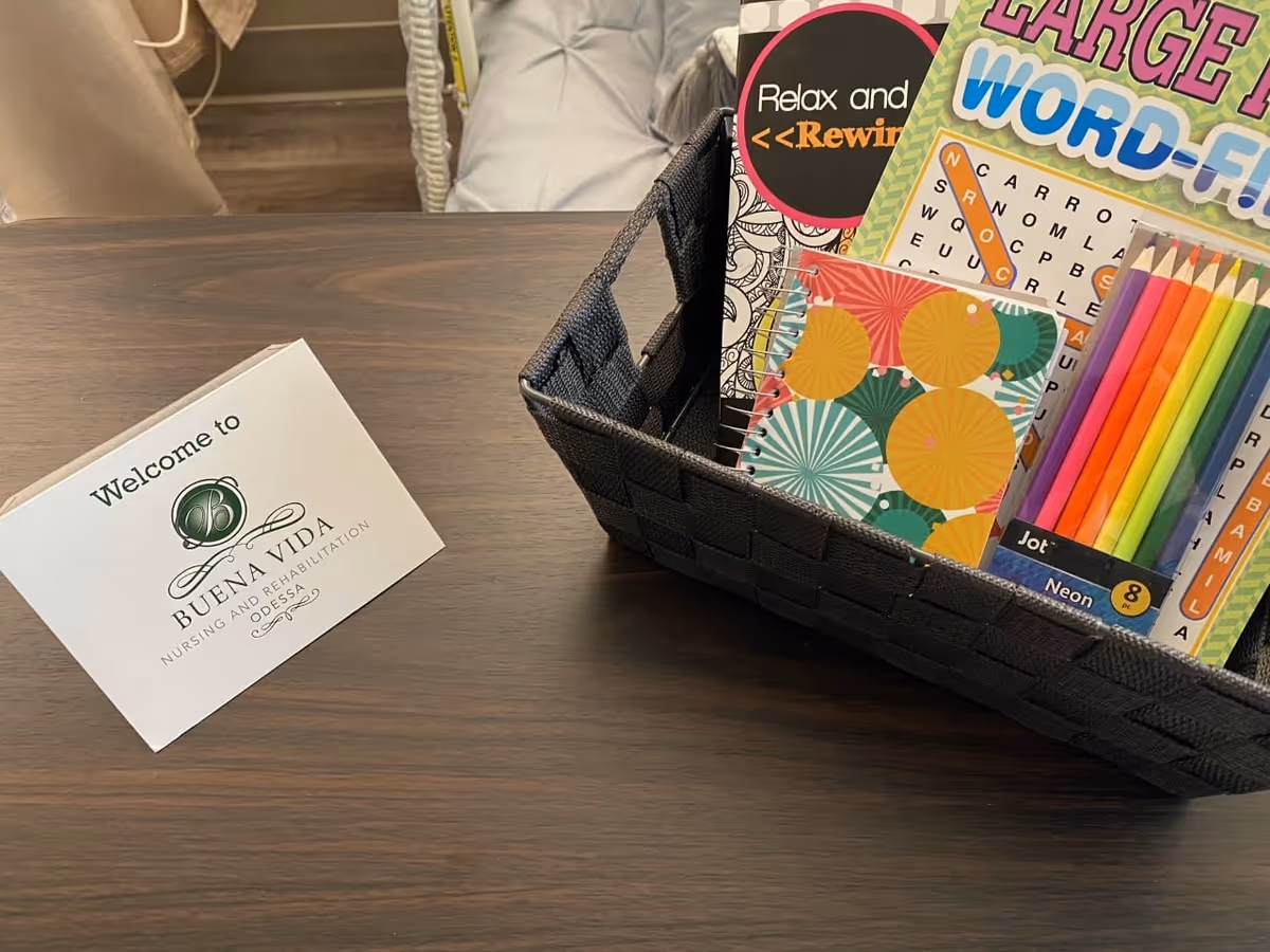 A wooden table with a welcome card from Buena Vida Nursing and Rehabilitation Odessa and a black woven basket containing a large print word-find puzzle book, colored pencils, a small colorful notebook, and a relaxation-themed booklet.