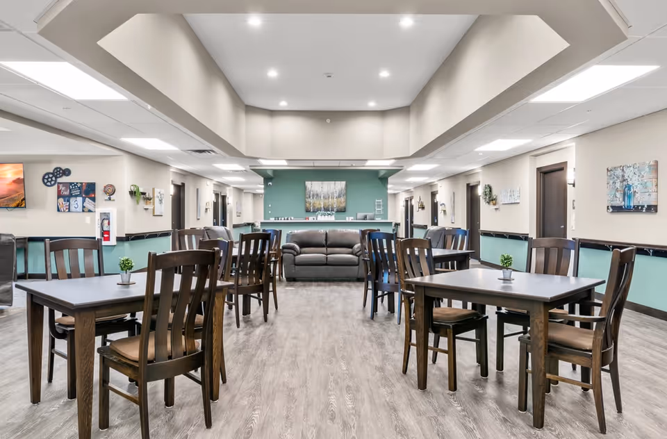 A spacious and well-lit common dining area in a senior living facility with multiple dark wooden tables and chairs arranged neatly. The room features light-colored walls with teal accents, a gray sofa against the far wall, decorative paintings, and ceiling lights providing bright illumination.