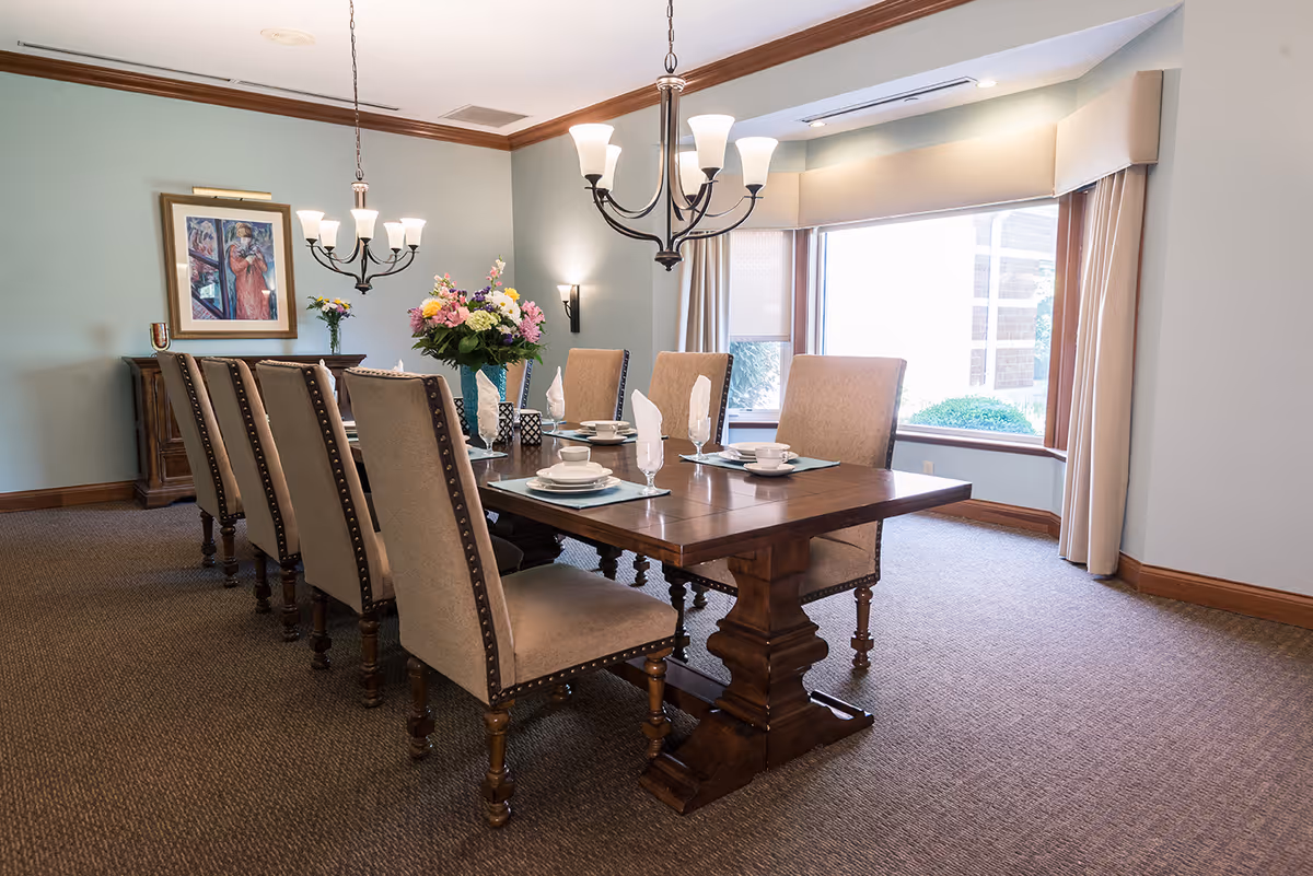 A formal dining room with a large wooden dining table set for six people. The table has beige upholstered chairs with nailhead trim, white napkins folded in glasses, white plates, and cups. A vase with colorful flowers is placed in the center of the table. The room has light blue walls, a large window with beige curtains, two chandeliers hanging from the ceiling, and a framed painting on the wall.