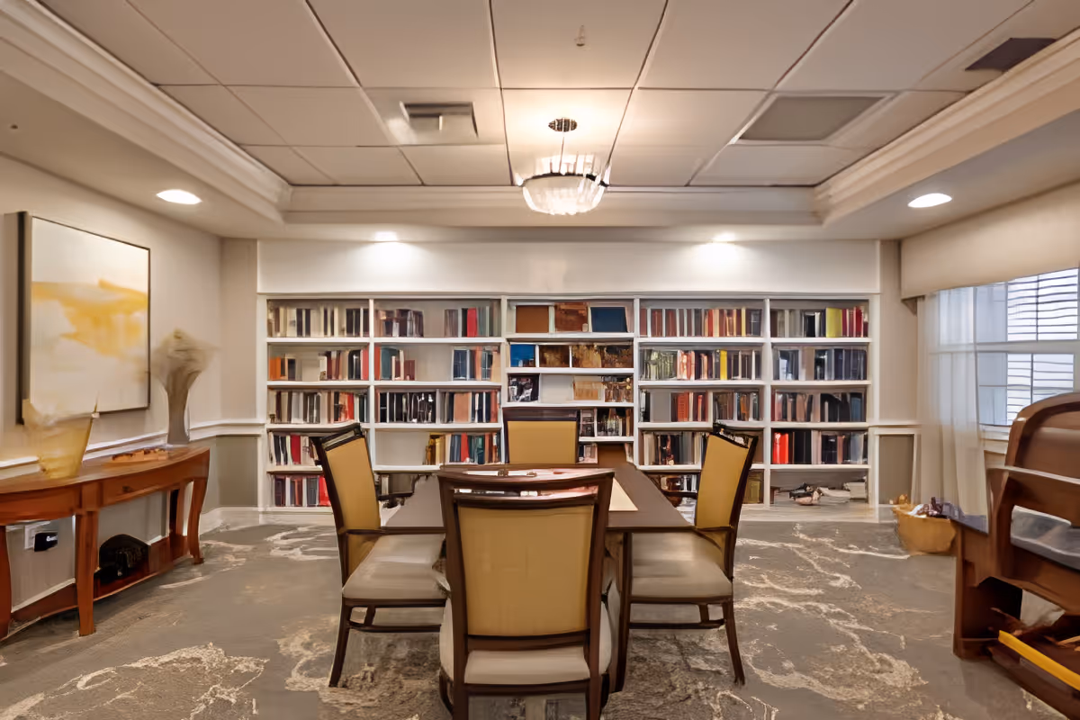 A cozy interior room with a rectangular wooden table surrounded by six cushioned chairs. Behind the table is a large built-in bookshelf filled with books. The room features a carpeted floor with a patterned design, a wooden side table with decorative items, a large window with sheer curtains, and a ceiling light fixture.