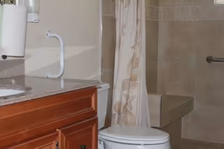Bathroom interior featuring a wooden vanity with a granite countertop and sink, a white toilet, a shower area with a beige patterned curtain, and tiled walls and floor with a built-in bench and a grab bar.