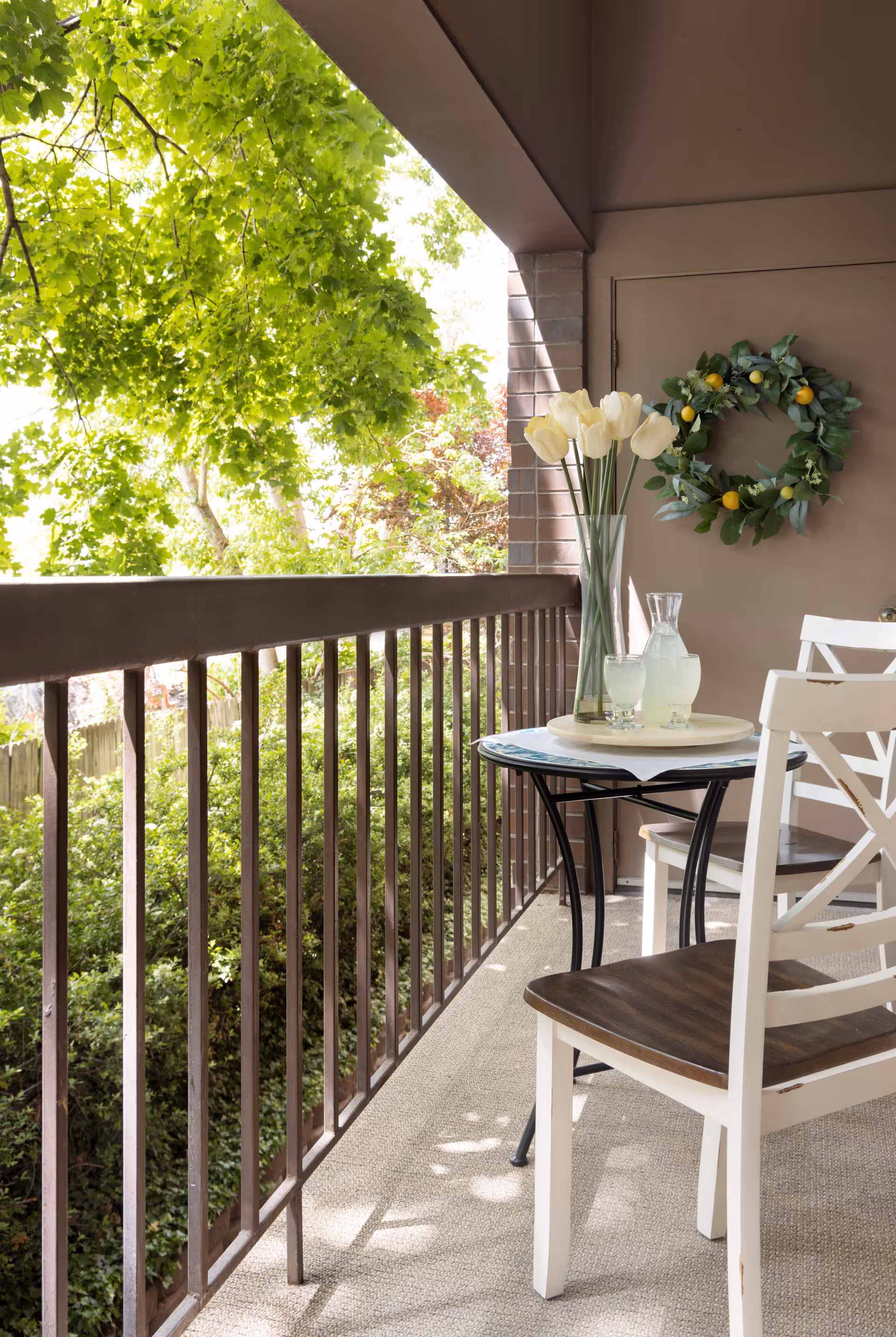A small outdoor balcony area with a round table and two white chairs with wooden seats. On the table, there is a vase with white tulips and a tray holding a glass pitcher and two glasses of lemonade. A green wreath with yellow accents hangs on the wall behind the table. The balcony overlooks lush green trees and bushes.