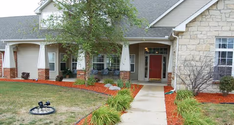 Front entrance of a residential building with a concrete walkway, landscaped mulch beds, and a covered porch with a red door.
