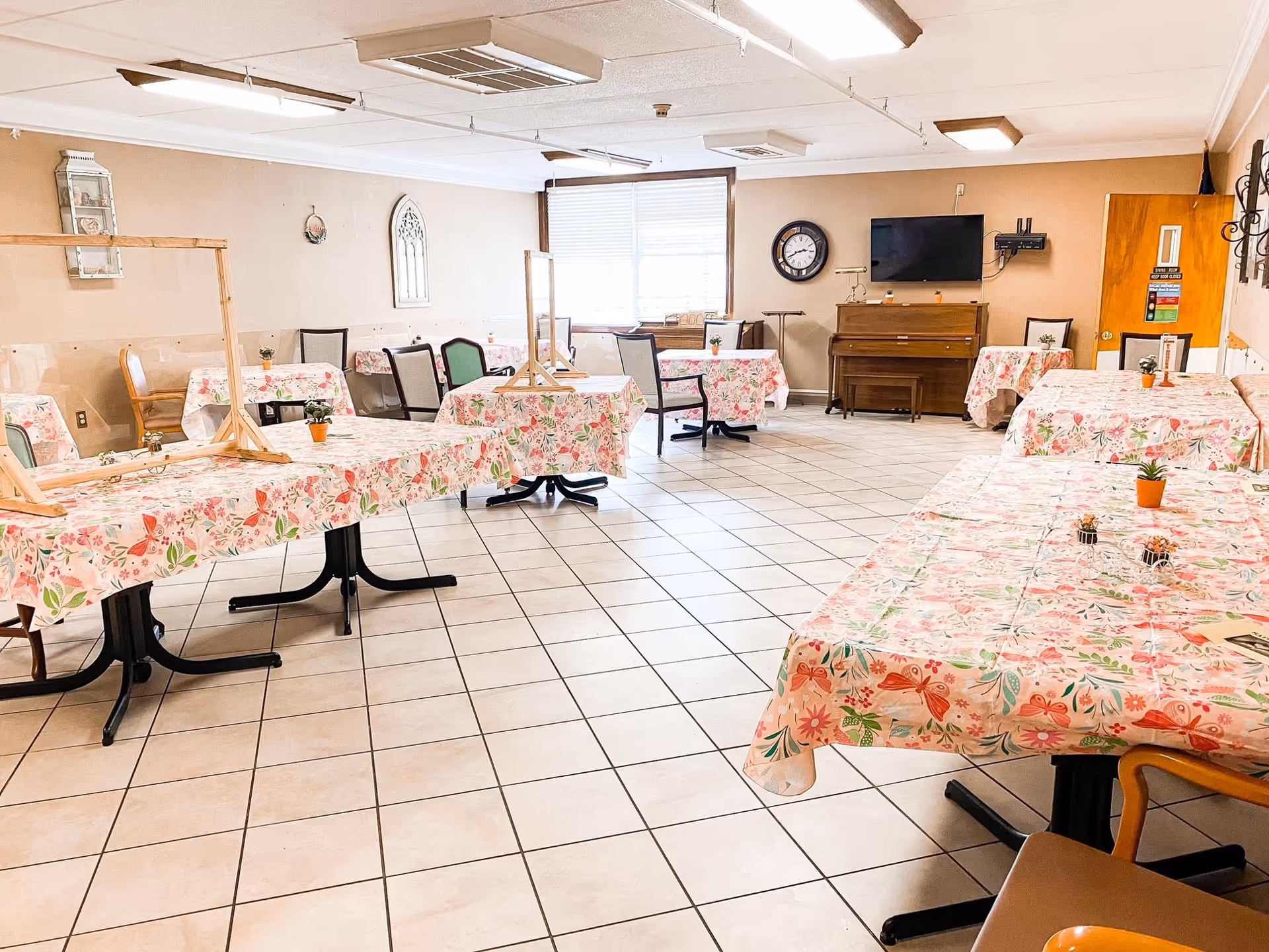 Empty communal dining/activity room with tiled floor, tables covered in floral tablecloths and plexiglass dividers, chairs, a piano and a wall-mounted TV.