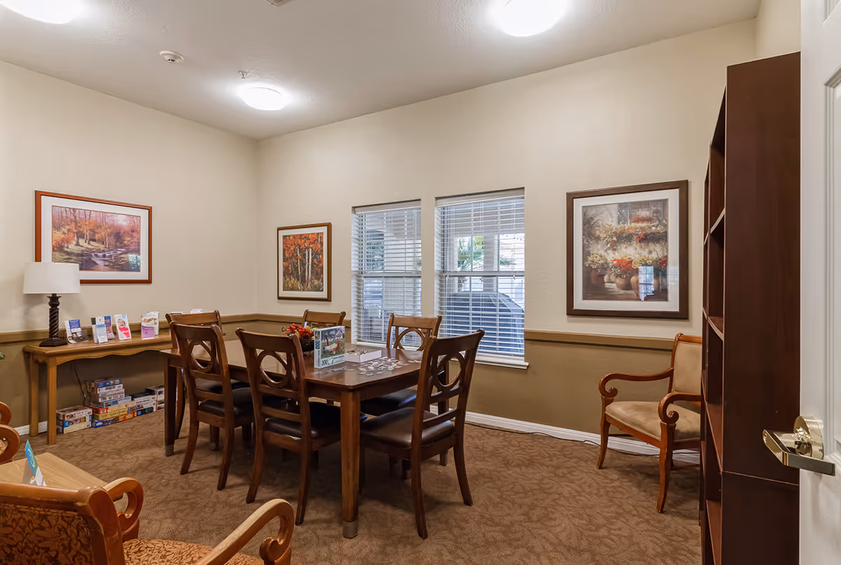 A cozy room with a wooden table surrounded by six chairs, a side table with a lamp and brochures, a bookshelf, and framed artwork on the walls. There are two windows with blinds letting in natural light, and a puzzle is partially completed on the table.