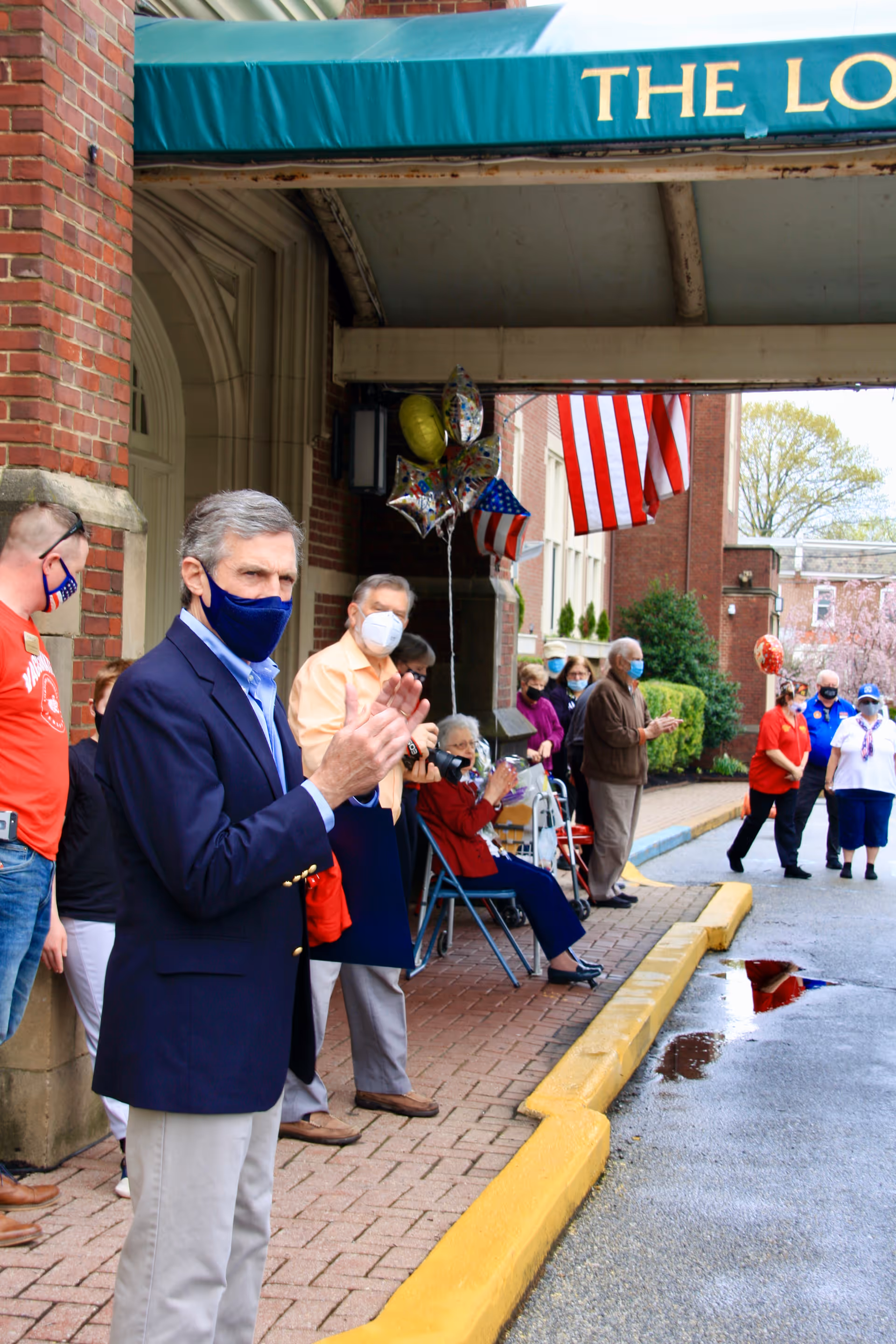A group of people, mostly elderly, standing and sitting outside a brick building under a green awning that reads 'THE LORELTON'. They are wearing face masks and clapping. There are American flags and balloons in the background.