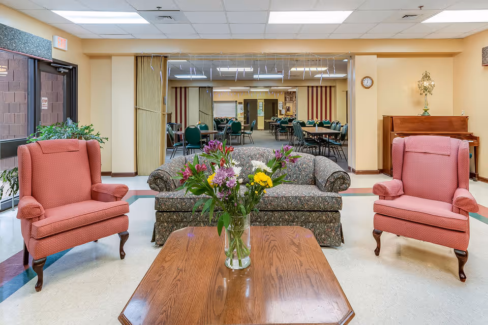 Seating area with two pink armchairs, a patterned sofa, and a coffee table topped with a vase of flowers opening into a dining room.
