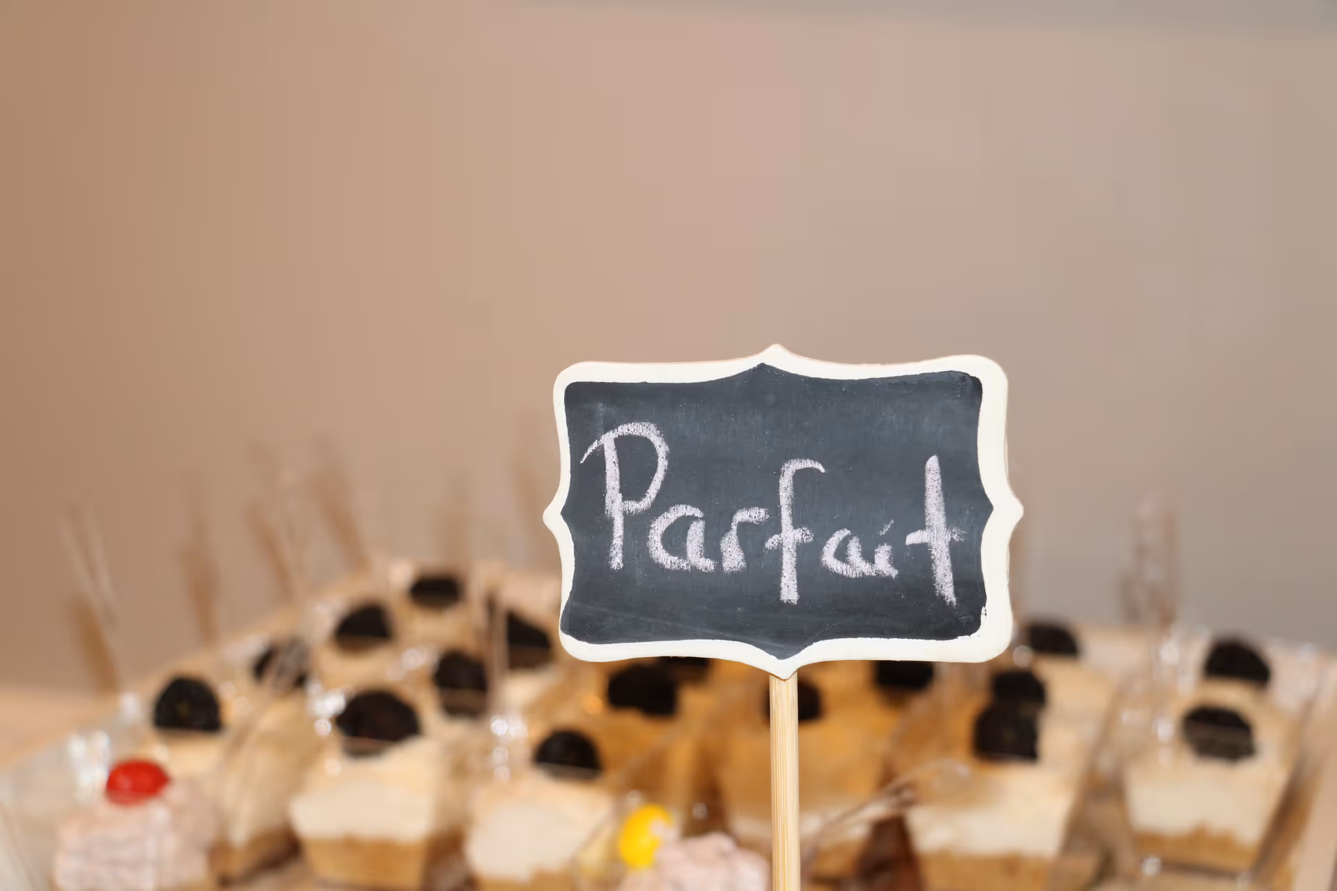 Close-up of a small chalkboard sign with the word 'Parfait' written on it, placed in front of several dessert parfaits in clear cups with spoons.